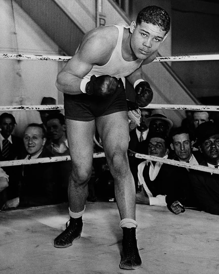 Black-and-white photograph of a boxer wearing gloves in a ring with spectators behind the ropes